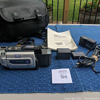Photo shows Sony Digital Handycam VR recorder with batteries, charger, cable, manual, and black leather carrying bag arranged on blue cloth surface.