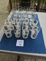 Two sets of glasses placed on a blue cloth. Front set of 8 glasses decorated with gold and black leaves. Back set of 12 clear glasses without color.