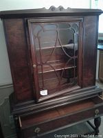 Front view of dark wood china cabinet with glass door and decorative wooden overlay showing three interior shelves and drawer below