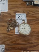 Pocket watch and accompanying chain necklace placed on wooden table with MaxSold tag