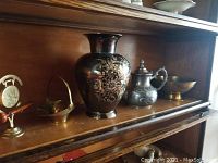 Wide view of shelf showing pewter teapot, brass bowl, brass urn, mini brass gong, brass basket, and brass stand with medallion.