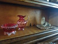 Wide shot of cranberry glass bowl and pitcher next to seal figurine on wooden stand inside a wooden cabinet.
