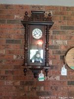 Front view of the wooden pendulum clock mounted on a brick wall, showing the clock face, carved wood details and pendulum through glass.