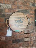 Wooden round wine cask lid with 'Chateau La Couspaude 2006 Grand Cru Classe Saint-Emilion' branding and rope handles hung on brick wall.