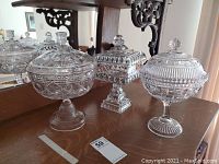 Three clear glass covered footed compotes on a table with intricate patterns and knobbed lids, includes two round and one square shaped compotes.