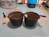 Two large cast iron pots side by side with visible rust and wear, placed on a basement floor.