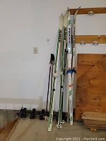 Photo showing two pairs of cross-country skis and poles leaning against a wall with a wooden rack nearby.