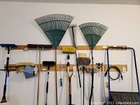 Front view of wall rack displaying brooms, rakes, brushes, hose, snow shovel, and garden hoe.