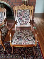 Full view of antique wooden rocking chair with brocade fabric on seat and back, positioned in front of brick fireplace on a patterned rug.