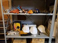 Shelving unit with three white hard hats, a boxed yellow and black trouble light, pairs of gloves, multiple extension cords in various colors and a beige power strip.