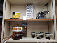 Shelf with pasta maker with metal body and black wooden handle, boxed pasta utensil, 6-piece fondue fork set, vintage floral enamel fondue pot on metal stand with wooden base, and two ceramic butter warmers with black metal stands.