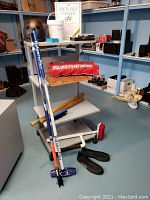 Photo of sports and games lot including baseball bats, curling broom handles, bocce balls, murder mystery game, and other equipment on floor and shelves.