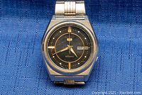 Front view of Seiko 5 automatic watch showing black dial with gold-tone markings and day-date window at 3 o'clock, minor scratches on crystal and case.