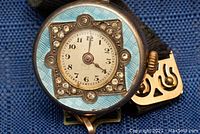 Front view of watch case showing light blue guilloché enamel, square bezel with clear stones and white Arabic numeral dial