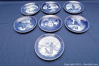 Seven blue and white Royal Copenhagen wall plates displayed on a blue cloth background, showing detailed seasonal and countryside scenes from 1972 to 1978.