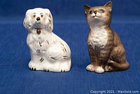 Front view of white dog figurine and brown and white cat figurine side by side on blue fabric background.