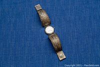 Watch laid flat showing entire band with embossed designs and round face with white dial and black numerals, on blue background.