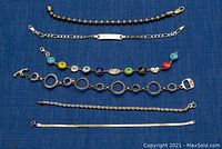 Six silver bracelets arranged on blue fabric, showing different styles and lengths including beaded, linked, flat snake chain and beaded charms.