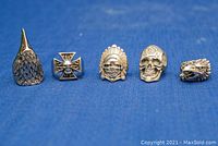 Front view of five large biker rings aligned on blue cloth showing detailed designs including bird head, iron cross with skull, Native American chief, skull with rose, and eagle head.