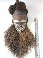 Front view of the mask showing wooden carving, painted face, dry grass beard, and fiber headdress with pom-poms, with ruler for scale.