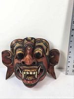 Front view of the Indonesian mask showing large fangs, prominent bulbous eyes, wide nose, and open mouth details with a measuring ruler beside it for scale.