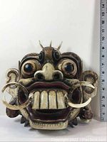 Front view of Indonesian wooden mask showing large bugged-out eyes, large fangs, and wooden earrings with size ruler on the side.