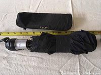 Black compact folded umbrella with silver and black handle and wrist strap, next to a tape measure showing about 12 inches in length.