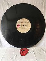 Photo of large 34-inch black vinyl record, showing label of The Rolling Stones with tongue logo and included fabric sew-on tongue logo patch beneath the record.