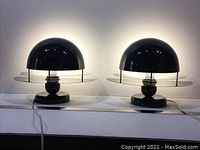 Two black lacquered metal dome lamps side by side on a white surface, illuminated; each with two smoked glass elliptical rings