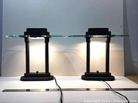 Pair of black dimmable table lamps with rectangular glass shelves shown side by side on a white surface with cords extended and lights turned on.