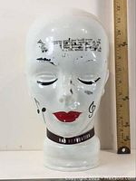 Front view of the full-size porcelain head with musical note designs on forehead, closed eyes, and red lips near a ruler showing approx. 12 inches in height.