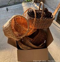 Photo of a box filled with five woven baskets of various sizes and shades, one basket contains pine cones, basket handle conditions vary including one loose handle.