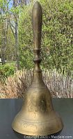 Brass bell photographed outdoors showing full view with handle, body engravings visible against natural background