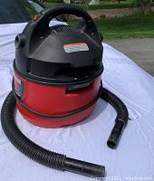 Photo of a small red and black Craftsman vacuum cleaner with a flexible hose attachment set on a white surface outdoors.