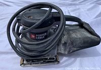 Small Porter Cable palm sander with power cord coiled on top, showing the rectangular sanding base and dust collection bag attached.