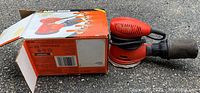Photo of Black and Decker palm sander sitting on pavement next to its original box, showing overall condition and model styling.