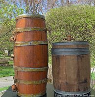 Photo showing two vintage wooden barrels outside on a table against trees and grass background. The taller barrel has several metal bands and metal handles, the shorter barrel has metal bands and a wooden spigot on top.