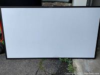 Photo of a large rectangular whiteboard placed on the ground outside, showing a clean white surface with a black border frame.