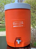 Orange Rubbermaid drinking water cooler with white lid and built-in spout, shown outside on a table with greenery in background.