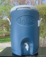 Front view of blue 5 gallon Igloo cooler with white spout and handles on sides, outdoor background with trees and grass.