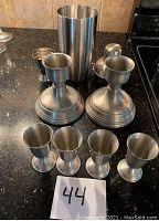Photo showing two large pewter candlestick holders, four small pewter apertifs, and one pewter shaker on a black countertop.