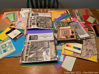 Photo showing a large collection of various sheets of colored and patterned scrapbook paper, three unopened kits, and assorted embellishments on a table.