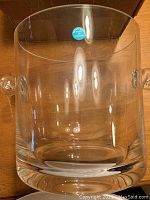 Clear glass champagne bucket with two handles and thick base, placed on wooden surface.