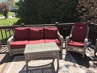Wicker outdoor couch with red cushions alongside wicker rocking chair with red cushion and matching wicker coffee table on wooden deck.