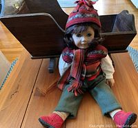 Photo of American Girl Doll Lindsey seated in front of a wooden doll cradle on a wooden floor. Doll wears red and gray striped hat and scarf, cream colored blouse, green pants, and red shoes.