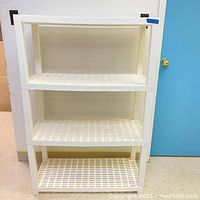 Front view of the white plastic storage shelf unit showing three lattice shelves and four corner support posts, placed on a tiled floor against a white wall and near a blue door.