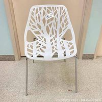 Front view of the white plastic chair with cutout tree branch pattern and chrome legs.