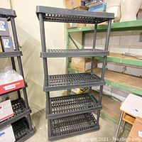 Black 5-level plastic storage shelf unit standing assembled in a warehouse setting.