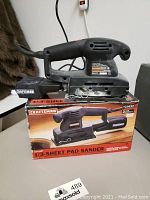 Front view of Craftsman 1/3 sheet pad sander sitting on top of its box showing the dust bag and power cord, with some cosmetic dust on the tool.