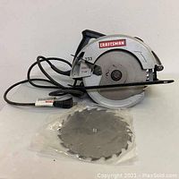 Side view of Craftsman circular saw showing blade guard, power cord, and extra blade in plastic packaging in front.